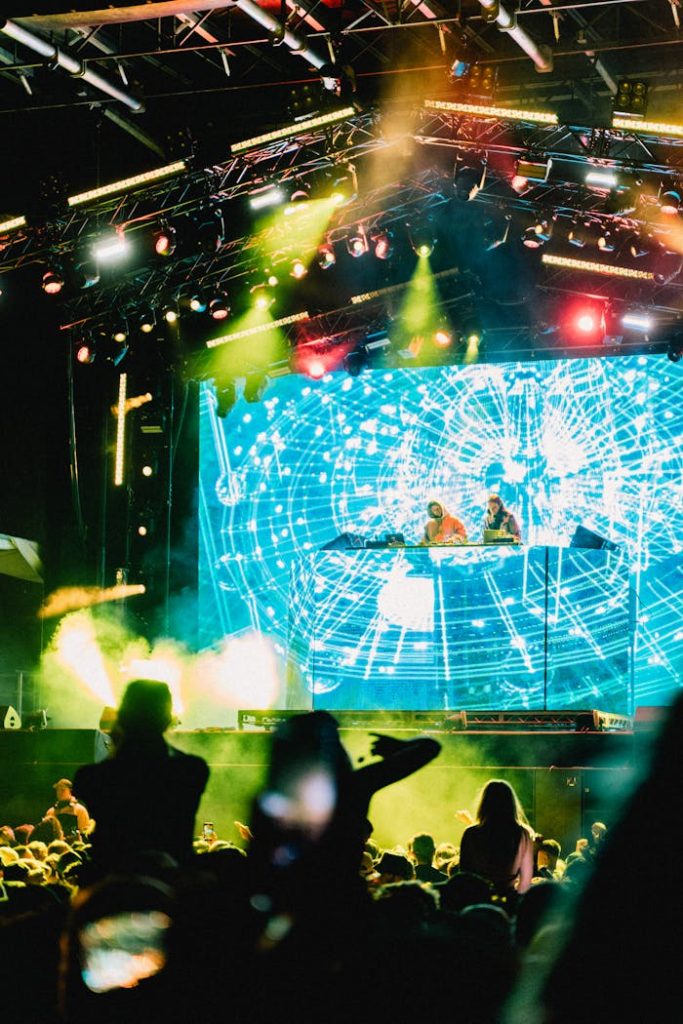Vibrant concert scene with DJs performing on stage amidst glowing lights and a cheering crowd.